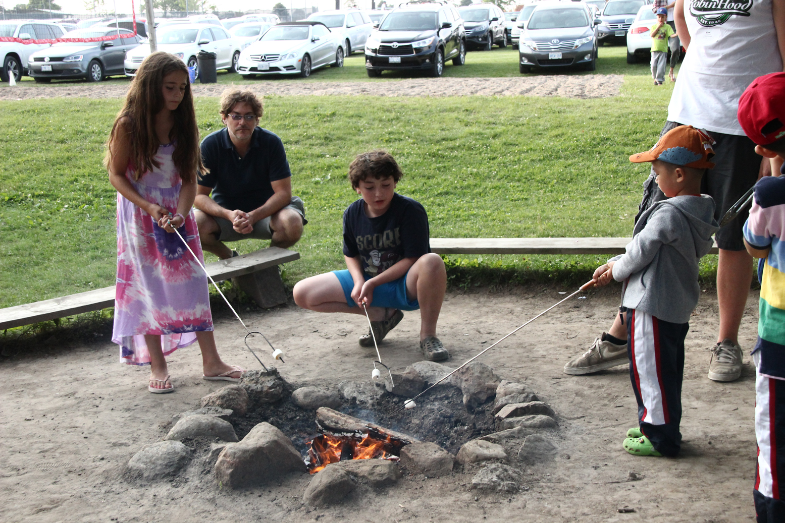 A girl and boy roast marshmallows on a long stick over a campfire at Camp Robin Hood. This camp provides traditional outdoor camp-style activities but is a day camp where children go home each night on free door-to-door bus service.